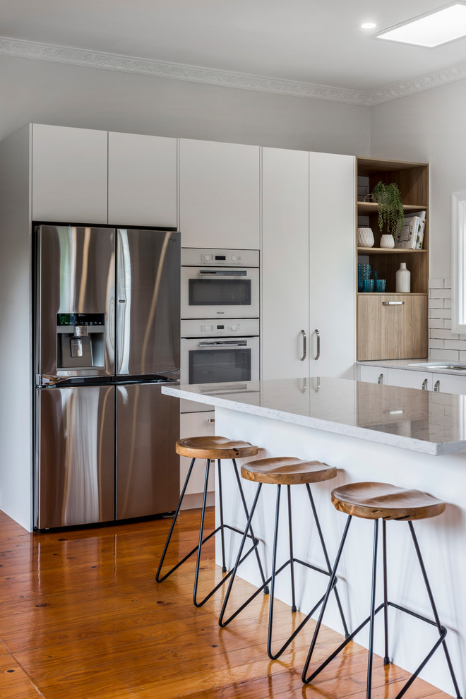Inspiration for a mid-sized contemporary l-shaped open plan kitchen in Brisbane with an undermount sink, flat-panel cabinets, white cabinets, quartz benchtops, white splashback, ceramic splashback, white appliances, medium hardwood floors, with island and brown floor.