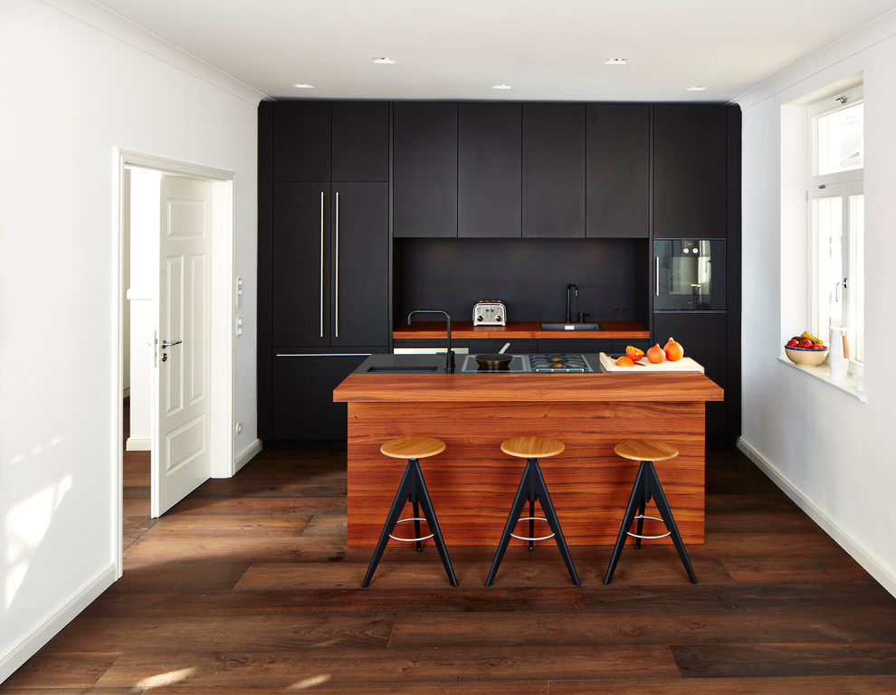 Small contemporary galley open plan kitchen in Munich with a drop-in sink, flat-panel cabinets, black cabinets, wood benchtops, black splashback, black appliances, dark hardwood floors and with island.