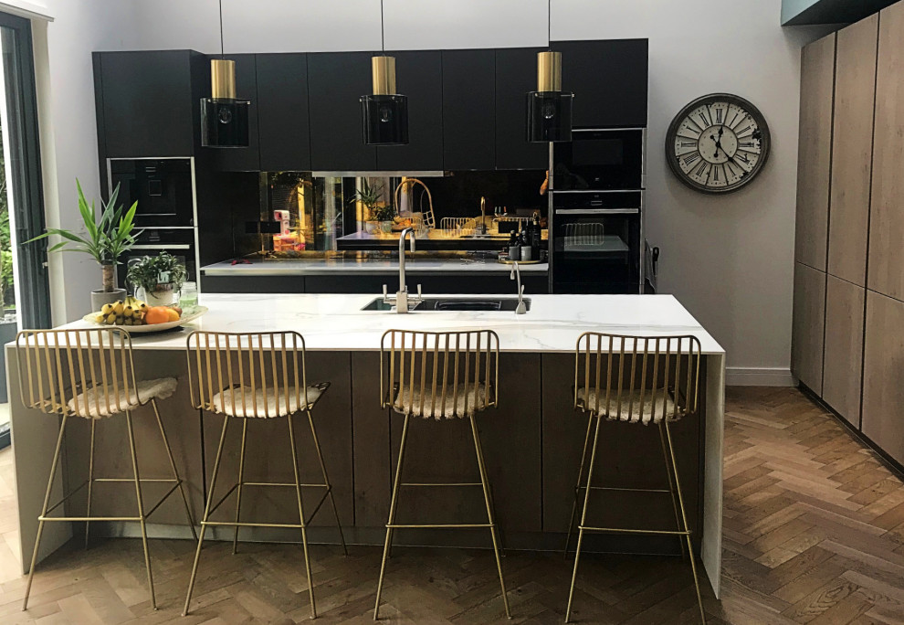 Photo of a contemporary open plan kitchen in Surrey with an undermount sink, flat-panel cabinets, black cabinets, solid surface benchtops, metallic splashback, glass sheet splashback, black appliances, plywood floors, with island, brown floor and white benchtop.