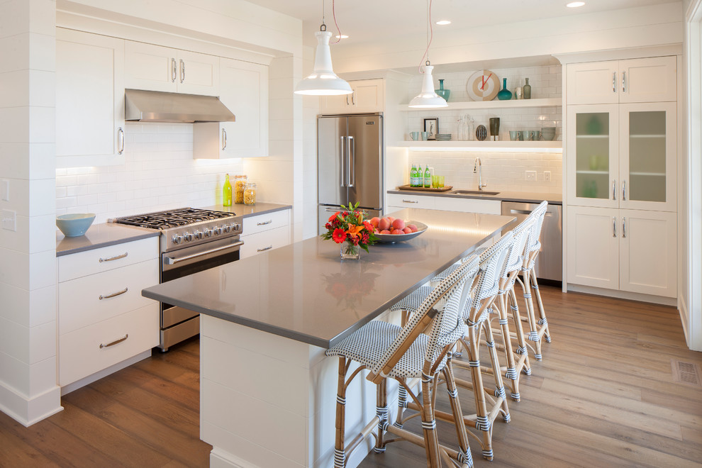 This is an example of a beach style l-shaped kitchen in Grand Rapids with an undermount sink, shaker cabinets, white cabinets, white splashback, subway tile splashback, stainless steel appliances, medium hardwood floors and with island.