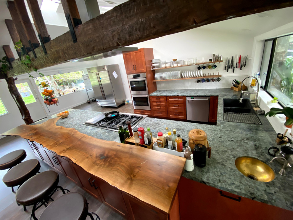 This is an example of a mid-sized beach style u-shaped eat-in kitchen in San Francisco with a drop-in sink, shaker cabinets, brown cabinets, granite benchtops, white splashback, ceramic splashback, stainless steel appliances, vinyl floors, a peninsula, brown floor, green benchtop and vaulted.