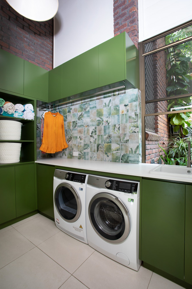 This is an example of a small modern l-shaped laundry room in Brisbane with a drop-in sink, green cabinets, quartz benchtops, green splashback, porcelain splashback, multi-coloured walls, porcelain floors, a side-by-side washer and dryer, grey floor and white benchtop.