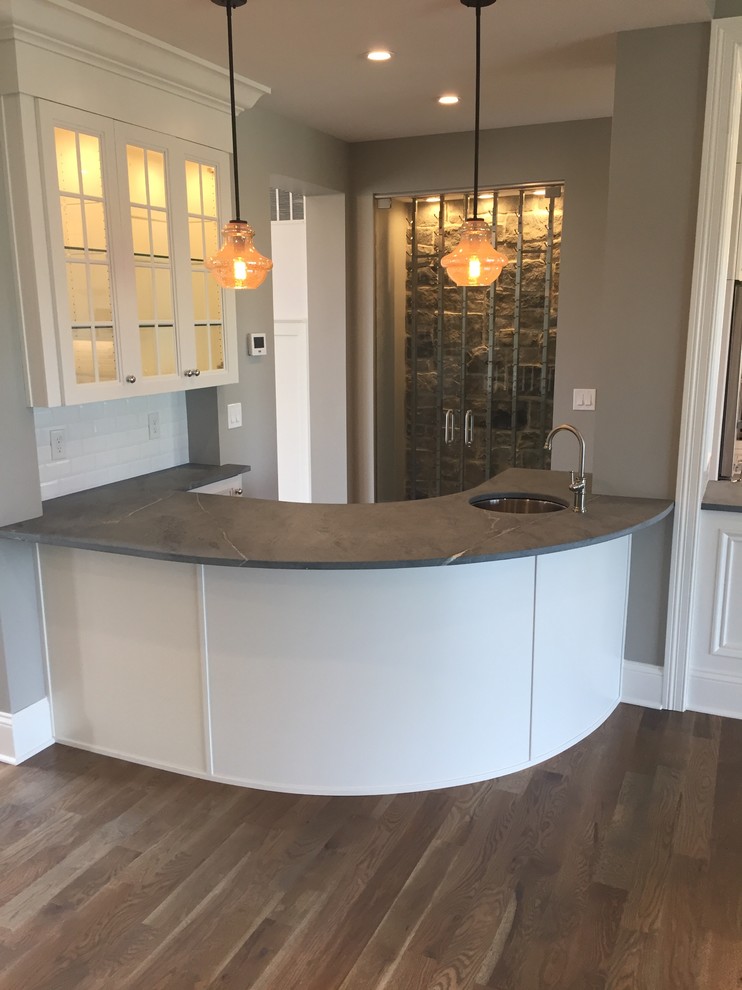 This is an example of a mid-sized transitional galley wet bar in Cincinnati with an undermount sink, glass-front cabinets, white cabinets, soapstone benchtops, white splashback, subway tile splashback, medium hardwood floors and grey floor.