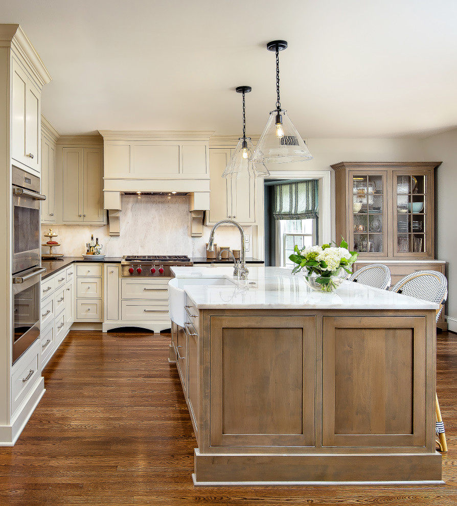 Inspiration for a large transitional l-shaped eat-in kitchen in Other with a farmhouse sink, shaker cabinets, granite benchtops, stainless steel appliances, medium hardwood floors, with island, black benchtop, beige cabinets, beige splashback, stone slab splashback and brown floor.