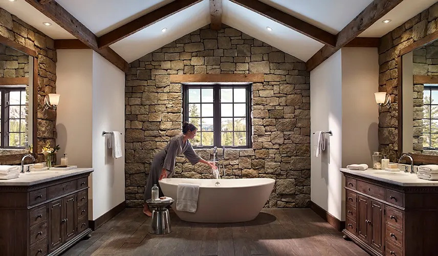 Country master bathroom in New York with furniture-like cabinets, dark wood cabinets, a freestanding tub, gray tile, stone tile, wood-look tile, brown floor, beige benchtops, a double vanity, a freestanding vanity, exposed beam and brick walls.