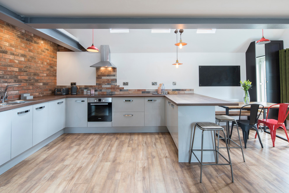 Mid-sized industrial u-shaped eat-in kitchen in West Midlands with a drop-in sink, flat-panel cabinets, grey cabinets, wood benchtops, brown splashback, medium hardwood floors, a peninsula, brown floor and brown benchtop.