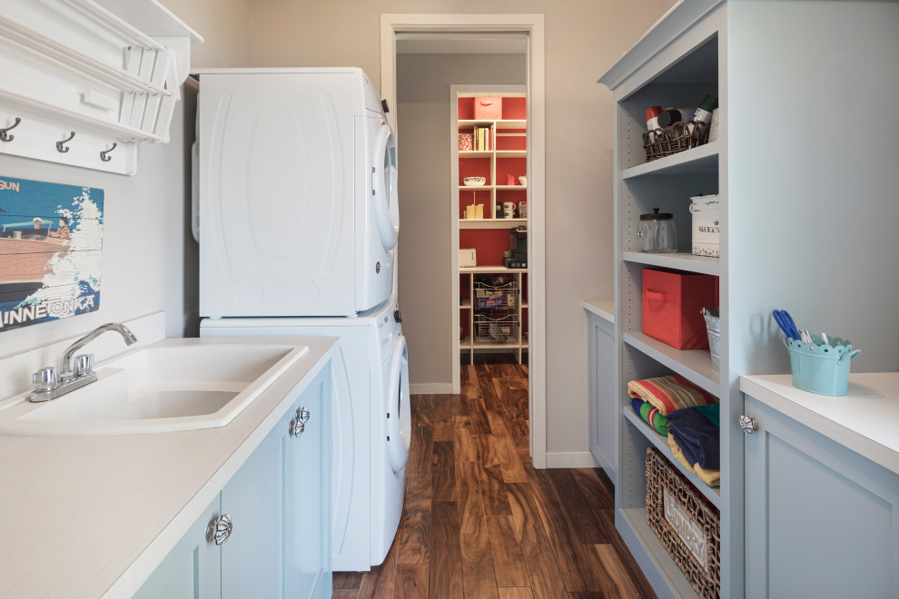Inspiration for a small beach style galley utility room in Minneapolis with a double-bowl sink, open cabinets, blue cabinets, grey walls, medium hardwood floors, a stacked washer and dryer and white benchtop.
