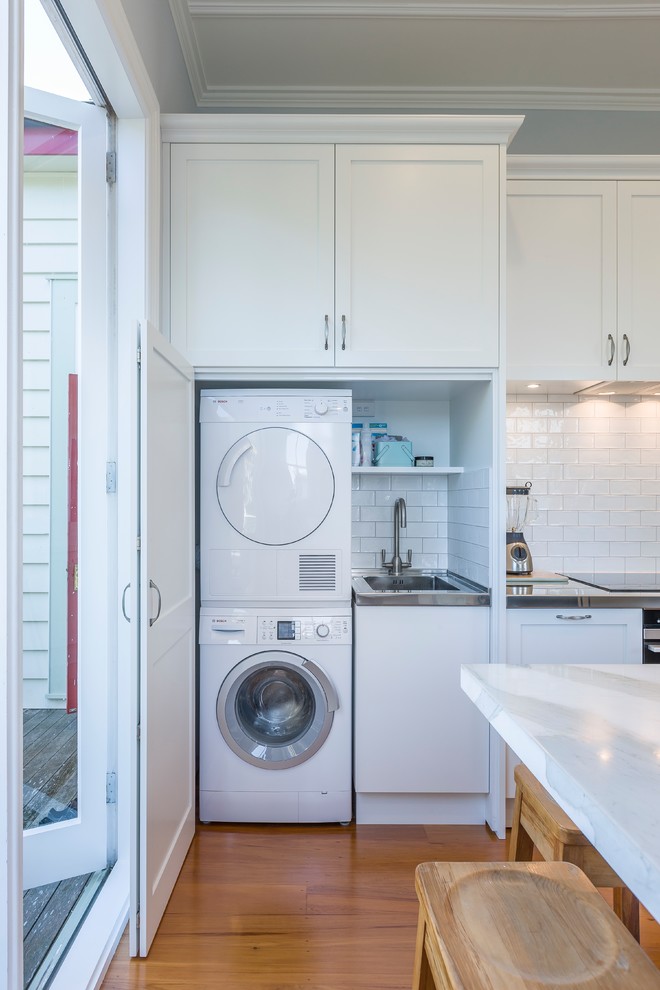 This is an example of a small single-wall utility room in Auckland with an utility sink, shaker cabinets, white cabinets, white walls, medium hardwood floors, a concealed washer and dryer and orange floor.