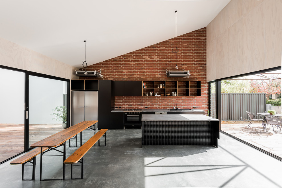 This is an example of an industrial single-wall open plan kitchen in Perth with black cabinets, concrete benchtops, brick splashback, stainless steel appliances, concrete floors, with island, grey floor, flat-panel cabinets and red splashback.