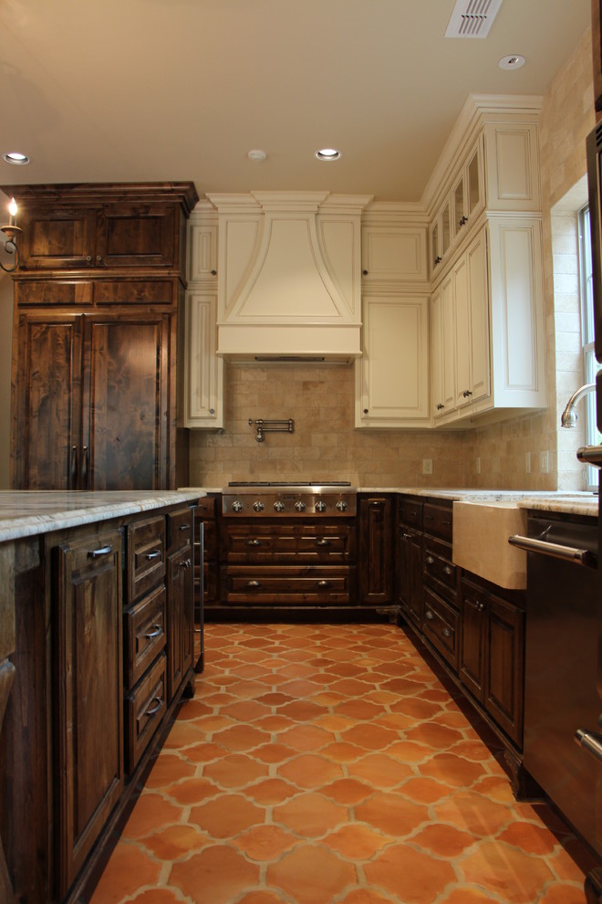 Photo of a large mediterranean l-shaped open plan kitchen in Dallas with a farmhouse sink, raised-panel cabinets, dark wood cabinets, granite benchtops, beige splashback, stone tile splashback, panelled appliances, terra-cotta floors and with island.