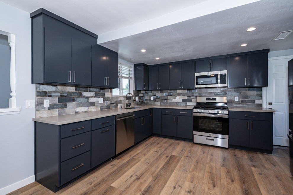 Large country l-shaped eat-in kitchen in Los Angeles with an undermount sink, flat-panel cabinets, blue cabinets, quartz benchtops, grey splashback, stone tile splashback, stainless steel appliances, light hardwood floors, no island, grey benchtop and brown floor.