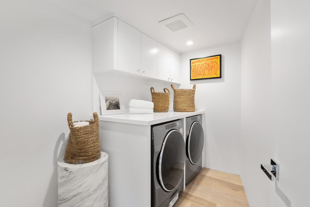 Photo of a contemporary single-wall dedicated laundry room in Los Angeles with flat-panel cabinets, white cabinets, white walls, light hardwood floors, a side-by-side washer and dryer, beige floor and white benchtop.