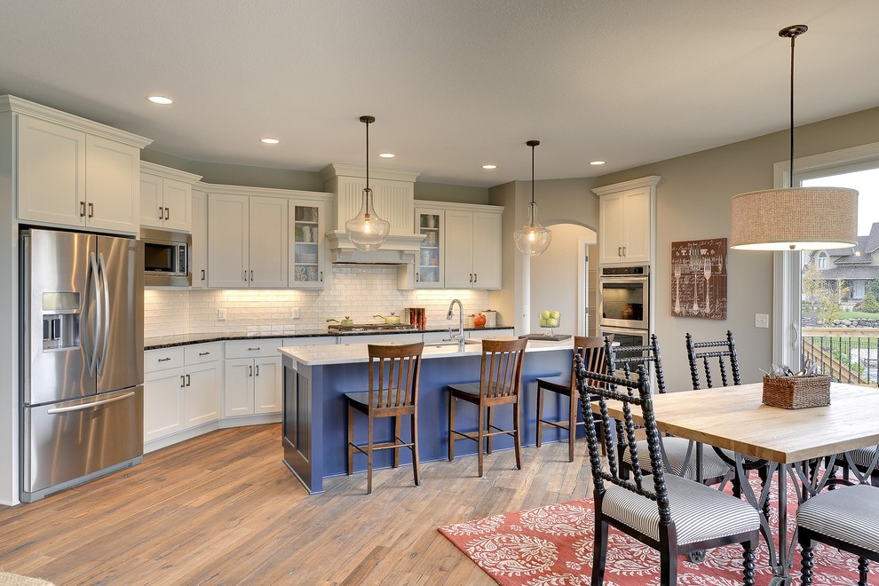 Photo of a large transitional single-wall eat-in kitchen in Minneapolis with a farmhouse sink, recessed-panel cabinets, white cabinets, white splashback, subway tile splashback, stainless steel appliances, medium hardwood floors and with island.