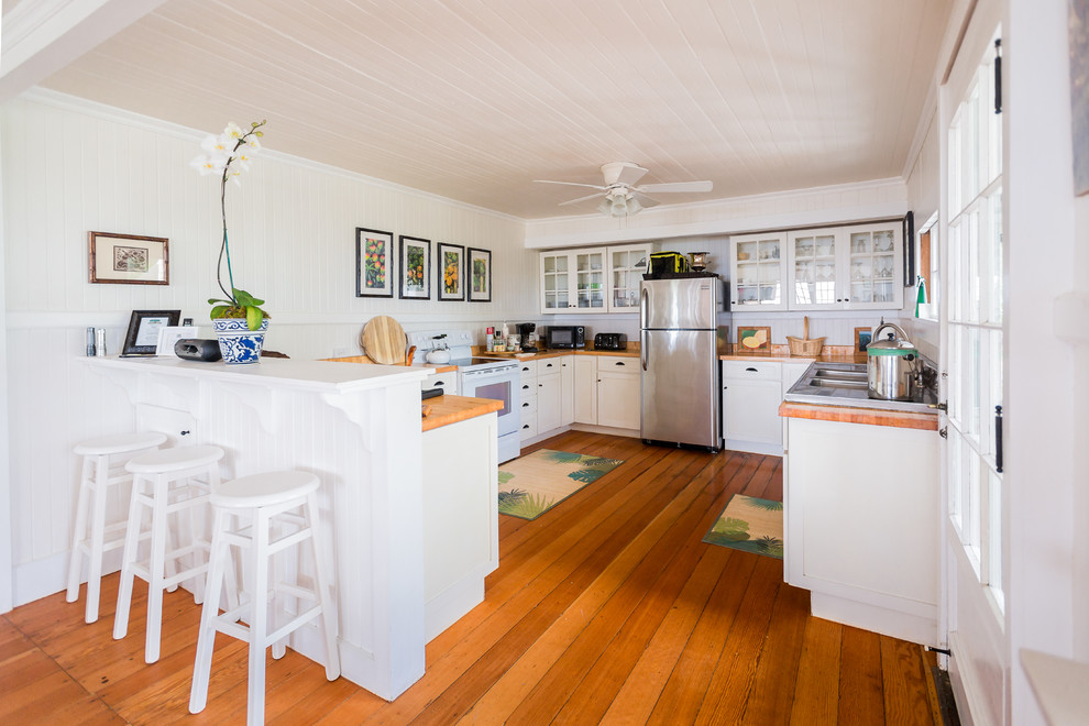 This is an example of a mid-sized tropical l-shaped eat-in kitchen in Hawaii with a double-bowl sink, flat-panel cabinets, white cabinets, wood benchtops, white splashback, stainless steel appliances, medium hardwood floors and a peninsula.