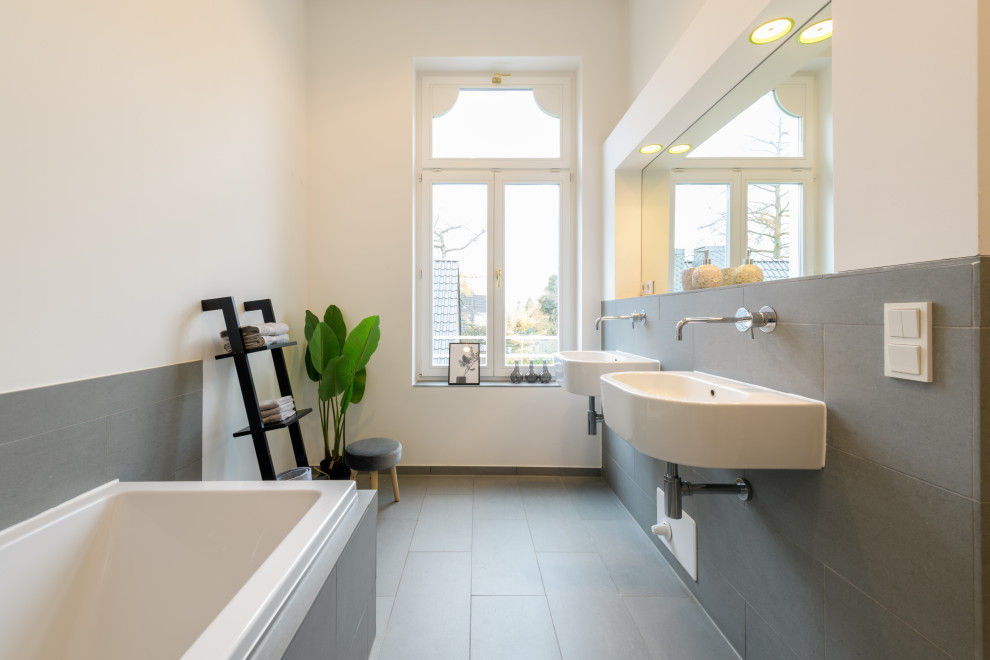 Esempio di una stanza da bagno minimal con vasca ad angolo, piastrelle grigie, pareti bianche, lavabo sospeso, pavimento grigio e due lavabi