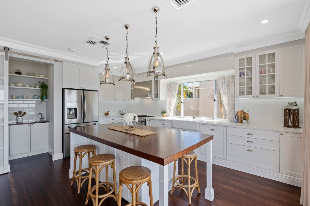 This is an example of a large transitional l-shaped eat-in kitchen in Gold Coast - Tweed with a farmhouse sink, shaker cabinets, white cabinets, wood benchtops, white splashback, porcelain splashback, stainless steel appliances, dark hardwood floors, with island, brown floor and brown benchtop.