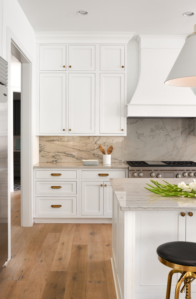 This is an example of a kitchen in New York with white cabinets, marble benchtops, beige splashback, marble splashback, stainless steel appliances, light hardwood floors, with island and beige benchtop.