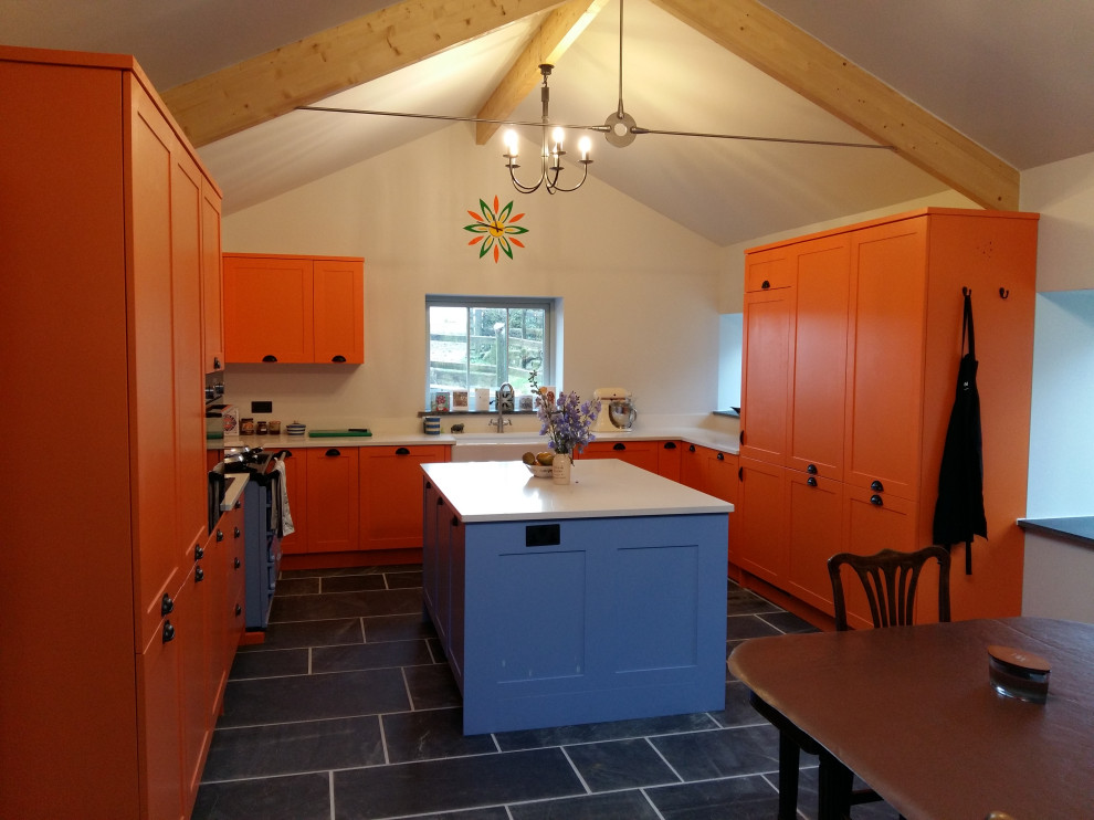 Inspiration for a large contemporary u-shaped open plan kitchen in Cornwall with a farmhouse sink, shaker cabinets, orange cabinets, quartz benchtops, white splashback, black appliances, slate floors, grey floor, white benchtop and exposed beam.