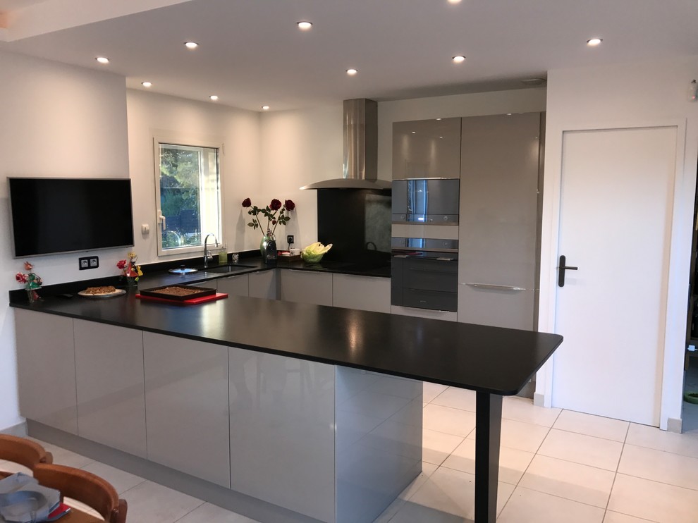 Cette photo montre cuisine américaine in U With un évier intégré, un placard à porte affleurante, des portes de placard grises, un plan de travail en granite, une crédence noire, un sol en carrelage de céramique, îlot, un sol beige and plan de travail noir.
