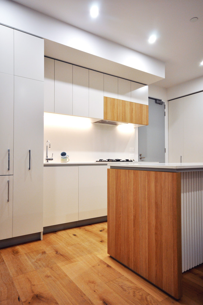This is an example of a medium sized modern single-wall open plan kitchen in Melbourne with a submerged sink, recessed-panel cabinets, medium wood cabinets, composite countertops, white splashback, stainless steel appliances, medium hardwood flooring, an island and white worktops.