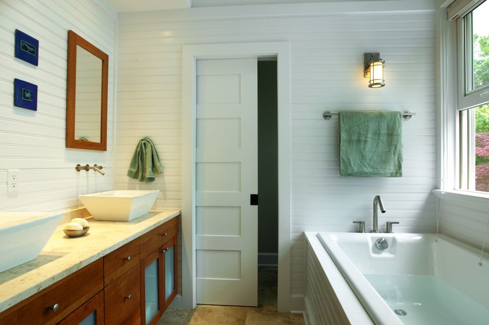 This is an example of a mid-sized beach style master bathroom in New York with shaker cabinets, medium wood cabinets, limestone benchtops, a drop-in tub, a vessel sink, white walls and travertine floors.