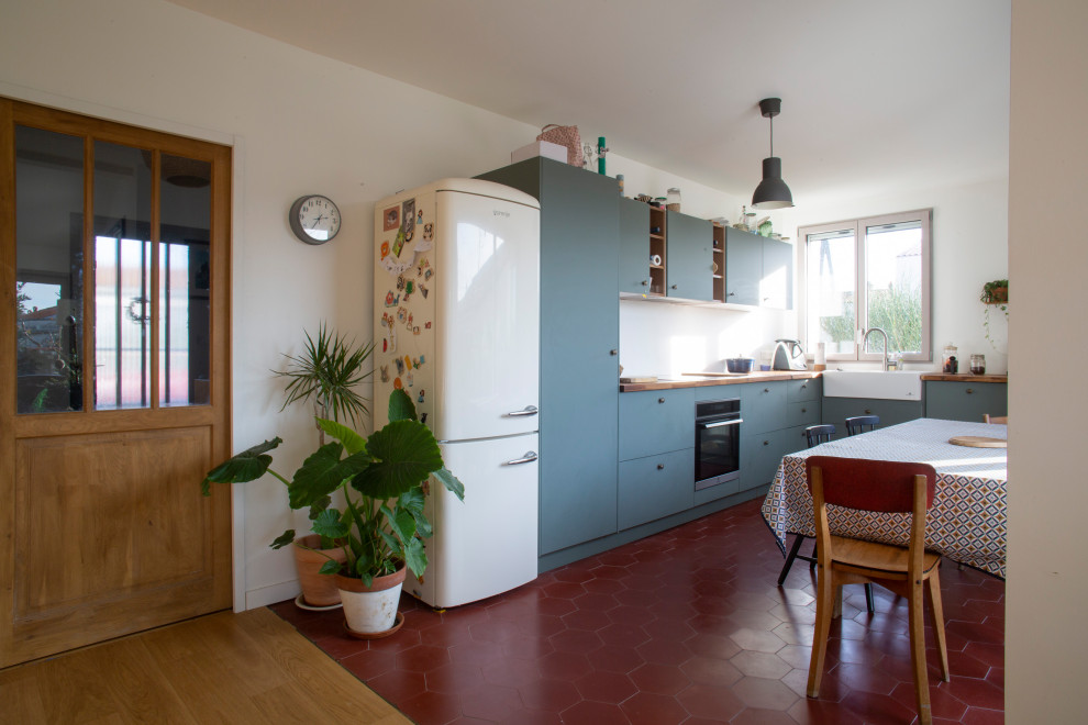 Photo of a mid-sized midcentury l-shaped open plan kitchen in Other with a drop-in sink, flat-panel cabinets, green cabinets, wood benchtops, black appliances, terra-cotta floors and red floor.