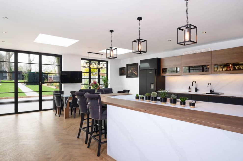 This is an example of a large industrial u-shaped open plan kitchen in Hertfordshire with an integrated sink, flat-panel cabinets, black cabinets, quartzite benchtops, white splashback, black appliances, light hardwood floors, with island, brown floor and white benchtop.