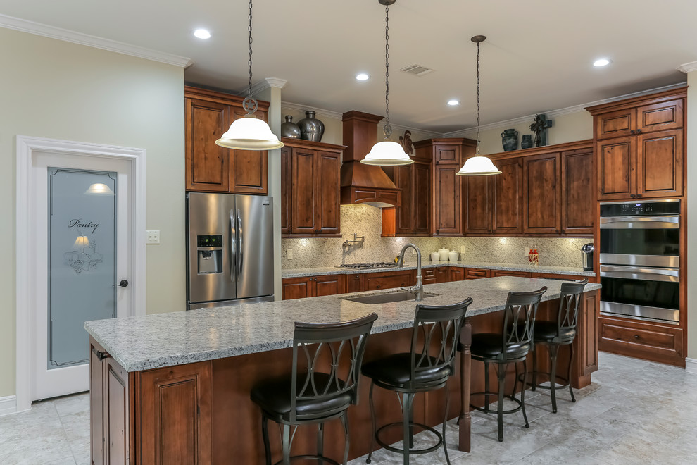 This is an example of a country l-shaped open plan kitchen in Houston with an undermount sink, raised-panel cabinets, medium wood cabinets, granite benchtops, grey splashback, stone slab splashback, stainless steel appliances, travertine floors and with island.
