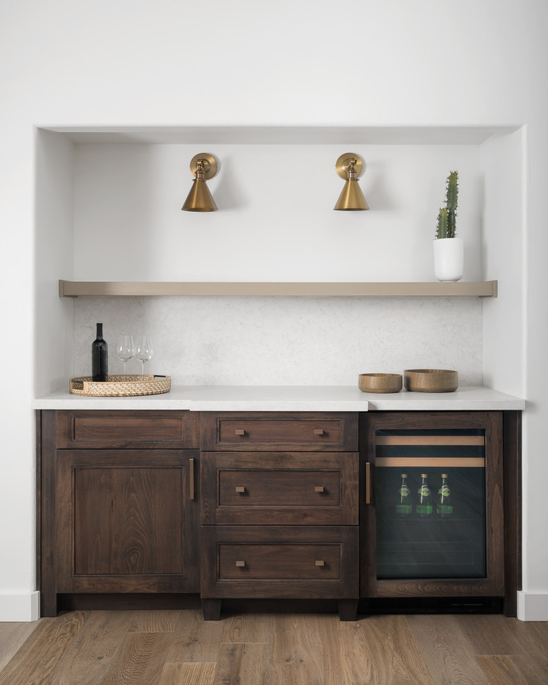 Photo of a small traditional single-wall wet bar in Phoenix with shaker cabinets, dark wood cabinets, white splashback, medium hardwood floors, brown floor and white benchtop.