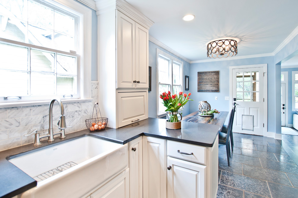 This is an example of a mid-sized transitional l-shaped eat-in kitchen in Nashville with a farmhouse sink, raised-panel cabinets, white cabinets, quartz benchtops, white splashback, subway tile splashback, stainless steel appliances, porcelain floors, with island, grey floor and black benchtop.