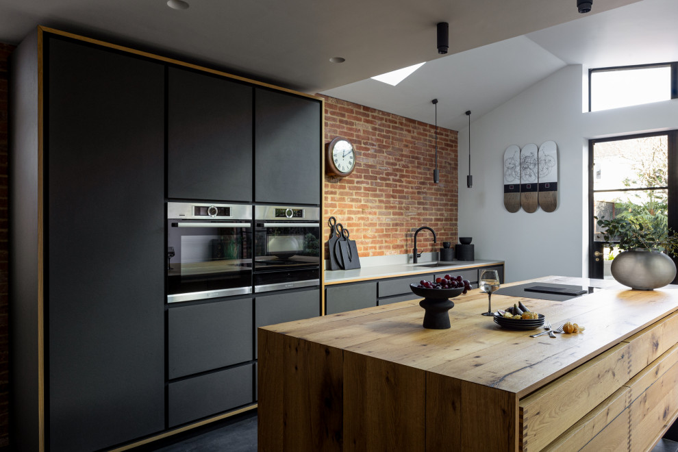 This is an example of a large contemporary grey and black single-wall open plan kitchen in Sussex with a built-in sink, flat-panel cabinets, distressed cabinets, quartz worktops, multi-coloured splashback, brick splashback, black appliances, ceramic flooring, an island, grey floors, grey worktops, a vaulted ceiling and a feature wall.