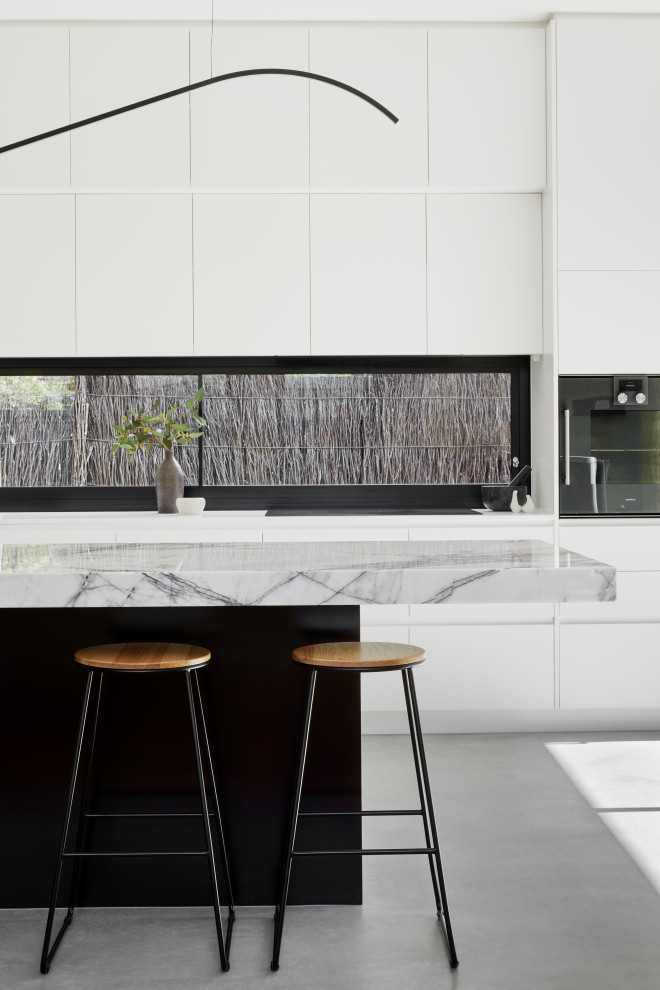 This is an example of a mid-sized contemporary galley open plan kitchen in Melbourne with an undermount sink, flat-panel cabinets, white cabinets, marble benchtops, black appliances, concrete floors, with island, grey floor and white benchtop.