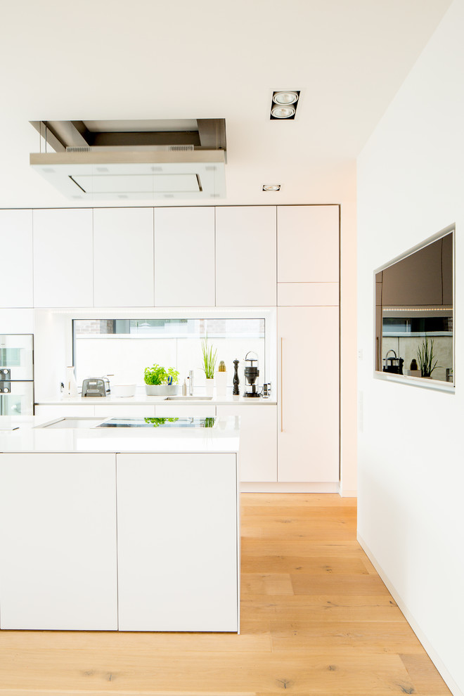 This is an example of a large modern galley open plan kitchen in Dusseldorf with an undermount sink, flat-panel cabinets, white cabinets, solid surface benchtops, white splashback, glass sheet splashback, white appliances, medium hardwood floors, with island, brown floor and white benchtop.