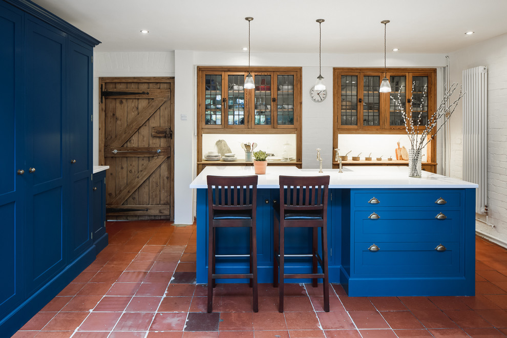 This is an example of a mid-sized country galley kitchen in Hertfordshire with a farmhouse sink, shaker cabinets, blue cabinets, terra-cotta floors, with island, red floor and white benchtop.