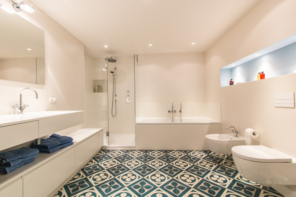 Photo of a large contemporary master bathroom in Hamburg with flat-panel cabinets, white cabinets, a drop-in tub, a wall-mount toilet, white tile, stone slab, white walls, cement tiles, an integrated sink, solid surface benchtops and blue floor.