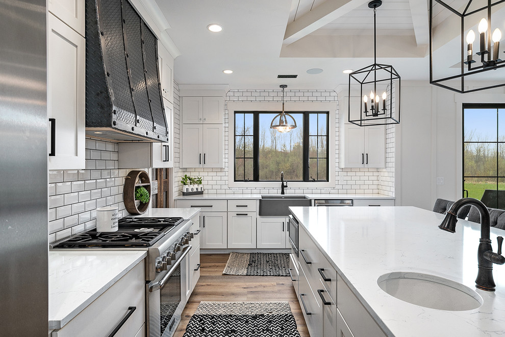 Inspiration for a country l-shaped kitchen pantry in Grand Rapids with a farmhouse sink, shaker cabinets, white cabinets, quartz benchtops, white splashback, ceramic splashback, stainless steel appliances, vinyl floors, with island, brown floor and white benchtop.