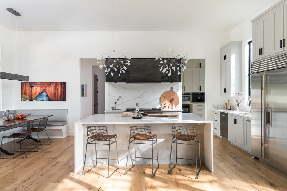 Photo of a contemporary u-shaped eat-in kitchen in Austin with shaker cabinets, grey cabinets, white splashback, stainless steel appliances, light hardwood floors, with island and brown floor.