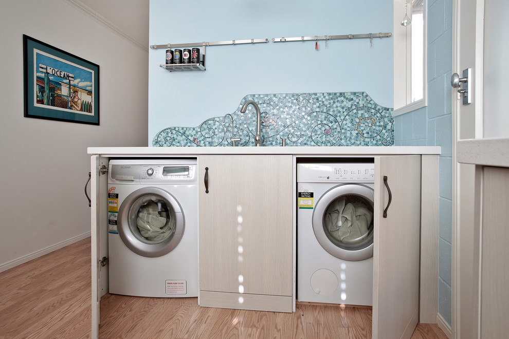 This is an example of a large contemporary single-wall utility room in Auckland with an undermount sink, flat-panel cabinets, beige cabinets, quartz benchtops, blue walls, laminate floors and a concealed washer and dryer.