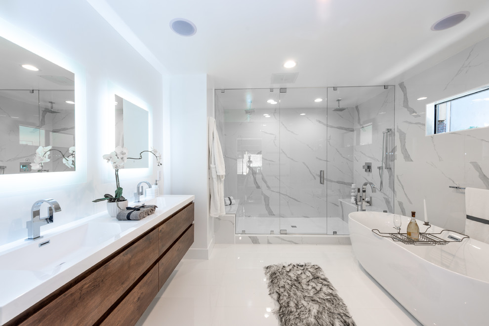 This is an example of a large contemporary master bathroom in Los Angeles with flat-panel cabinets, brown cabinets, a freestanding tub, a double shower, a two-piece toilet, white tile, porcelain tile, white walls, porcelain floors, an integrated sink, engineered quartz benchtops, white floor, a hinged shower door and white benchtops.