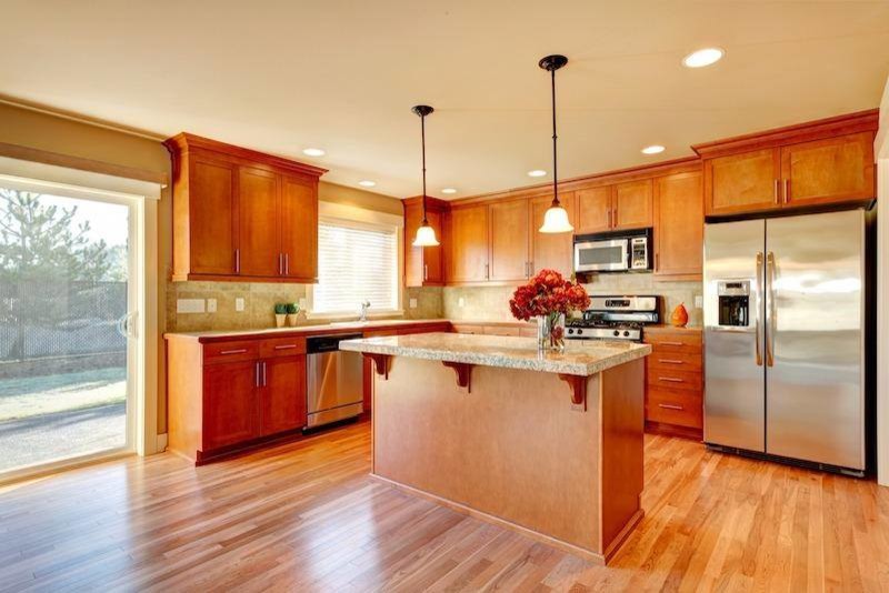 This is an example of a mid-sized traditional l-shaped open plan kitchen in Detroit with a drop-in sink, shaker cabinets, light wood cabinets, granite benchtops, beige splashback, stone tile splashback, stainless steel appliances, light hardwood floors, with island, beige floor and beige benchtop.