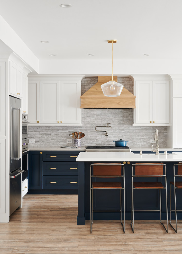 Design ideas for a large beach style l-shaped open plan kitchen in San Francisco with a farmhouse sink, shaker cabinets, white cabinets, solid surface benchtops, grey splashback, stainless steel appliances, light hardwood floors, with island, brown floor and white benchtop.