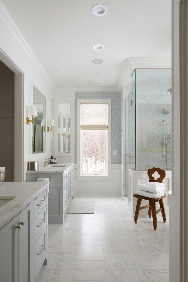 This is an example of a large classic ensuite bathroom in Minneapolis with recessed-panel cabinets, blue cabinets, a freestanding bath, an alcove shower, a two-piece toilet, white tiles, marble tiles, multi-coloured walls, marble flooring, a built-in sink, marble worktops, white floors, a hinged door, white worktops, a shower bench, double sinks, a built in vanity unit and wallpapered walls.
