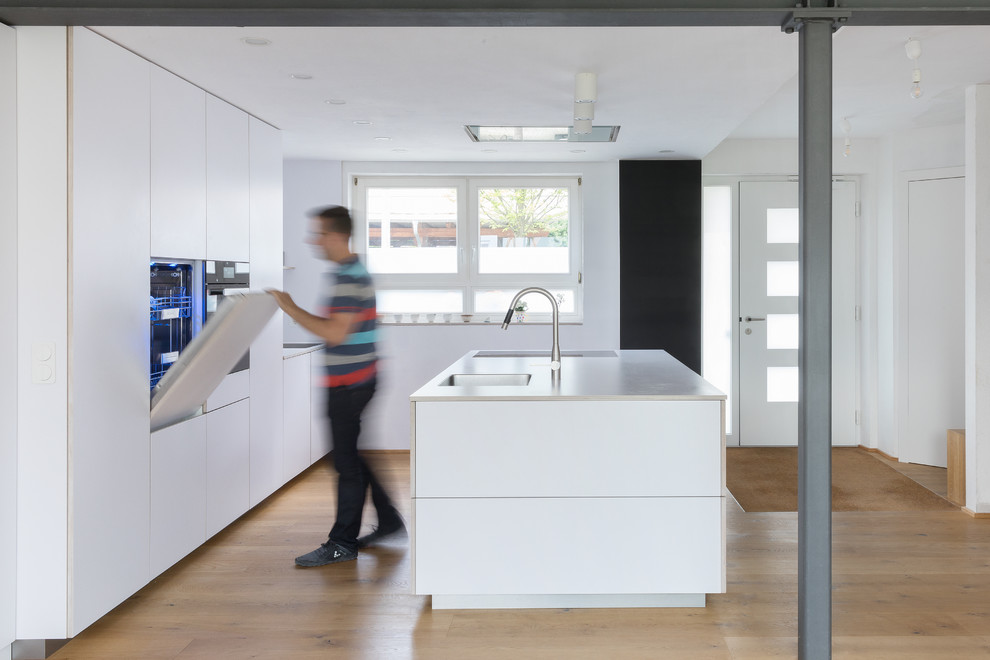 Inspiration for a mid-sized modern single-wall eat-in kitchen in Stuttgart with an undermount sink, white cabinets, laminate benchtops, white splashback, black appliances, light hardwood floors, with island, brown floor, white benchtop and flat-panel cabinets.