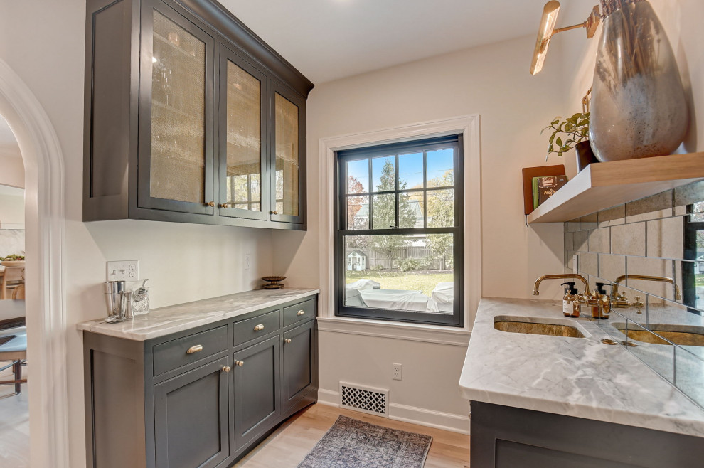 Photo of a small galley wet bar in Milwaukee with an undermount sink, shaker cabinets, black cabinets, marble benchtops, mirror splashback, light hardwood floors, brown floor and white benchtop.