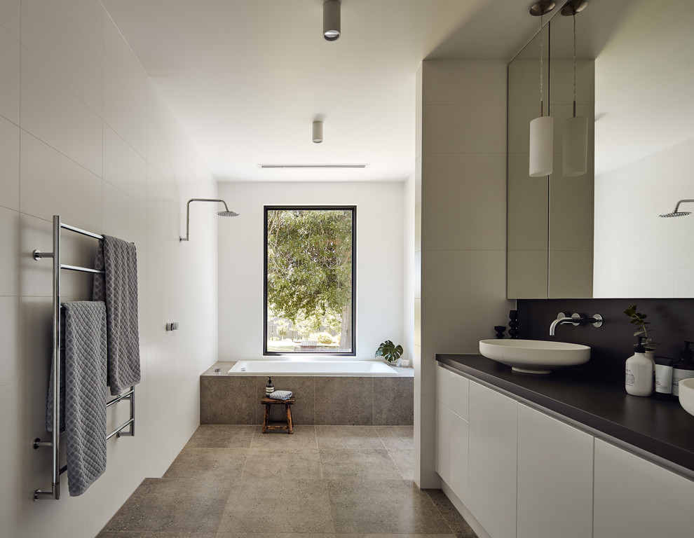 This is an example of a large contemporary master bathroom in Melbourne with a drop-in tub, an open shower, a one-piece toilet, white tile, ceramic tile, white walls, porcelain floors, a vessel sink, grey floor, an open shower and black benchtops.