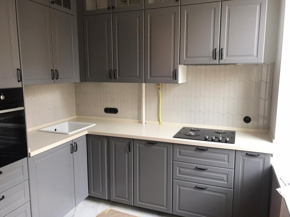 Photo of a small contemporary l-shaped separate kitchen in Moscow with a drop-in sink, raised-panel cabinets, grey cabinets, solid surface benchtops, white splashback, ceramic splashback, black appliances, no island and white benchtop.