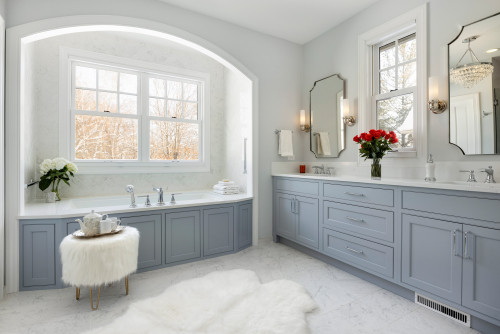 Elegant bathroom with light blue shaker-style bathroom cabinets, double sinks, marble countertops, and built-in bathtub under arched window with natural light