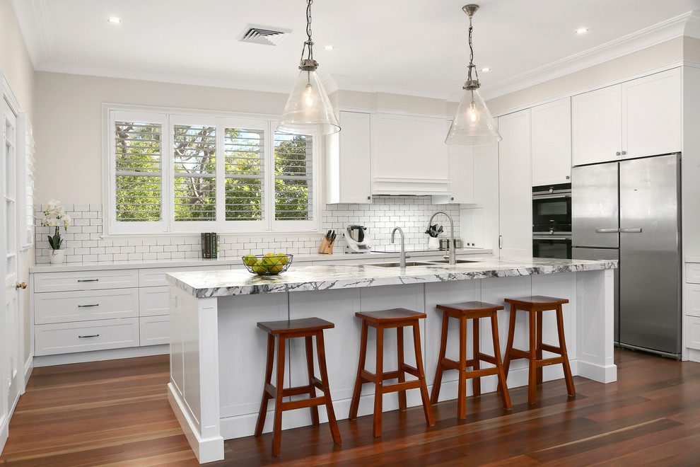 Inspiration for a transitional u-shaped kitchen in Newcastle - Maitland with an undermount sink, shaker cabinets, white cabinets, white splashback, subway tile splashback, stainless steel appliances, medium hardwood floors, with island, brown floor and white benchtop.
