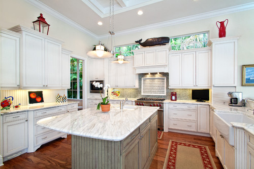 Bright traditional kitchen with white kitchen cabinets, marble island, farmhouse sink, and warm wood floors accented by decorative red accessories.