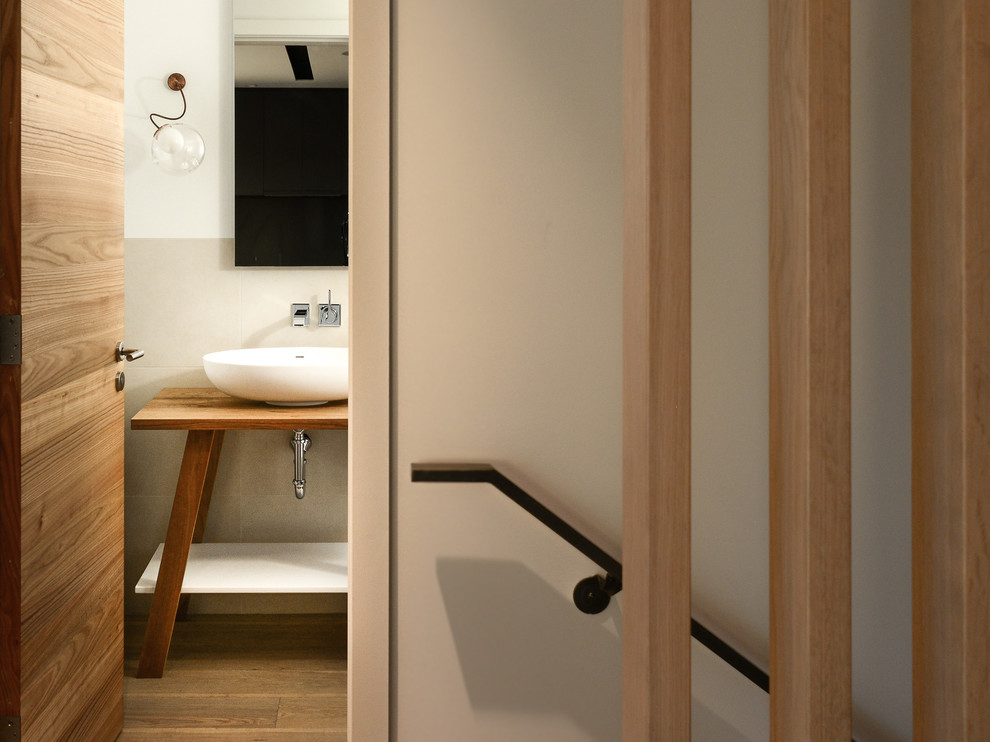 This is an example of a mid-sized modern powder room in Vancouver with light wood cabinets, a two-piece toilet, beige tile, porcelain tile, white walls, porcelain floors, furniture-like cabinets, a vessel sink, wood benchtops and brown benchtops.